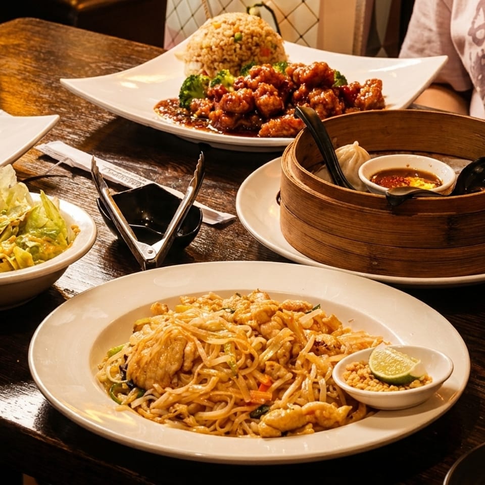 A table laden with various Asian dishes, including Pad Thai, orange chicken, salad, and dim sum.